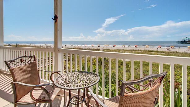 A balcony with wicker chairs and a table, overlooking a beach with umbrellas and a pier. - Seaside Villas II, Oceanfront Condo (Folly Beach)