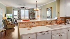 A modern kitchen with a central island, a dining area with a view, and a living room with a sofa and TV. - Seaside Villas II, Oceanfront Condo (Folly Beach)