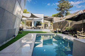 Uma área de piscina moderna ao ar livre, com design geométrico, cercada por um terraço com assentos e plantas em vasos.