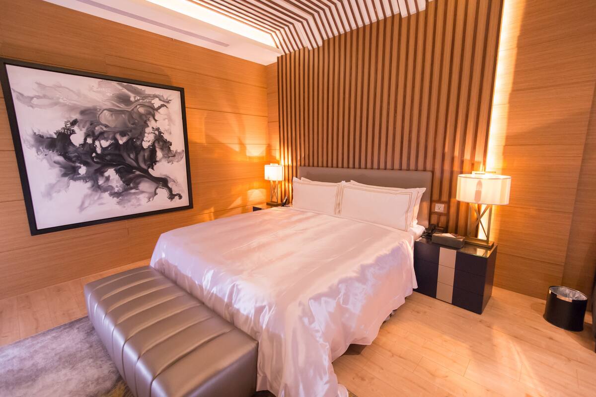 Ein modernes Hotelzimmer mit einem großen Bett, einer Bank, zwei Nachttischen mit Lampen und einem gerahmten abstrakten Gemälde an der Wand.