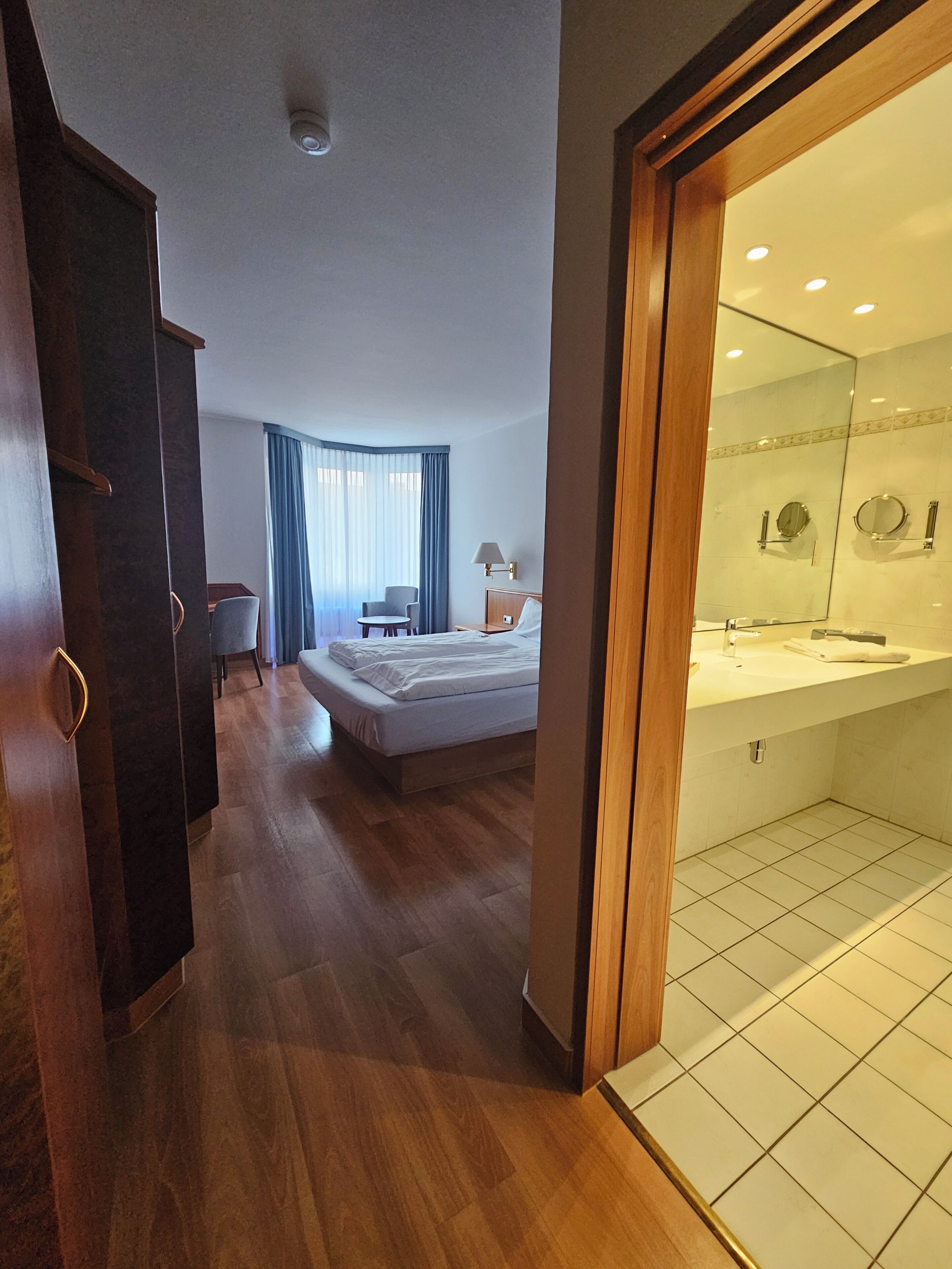 A hotel room with a bed, a desk, a chair, a wardrobe, and a bathroom with a mirror and sink.