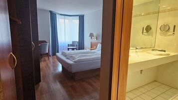 A hotel room with a bed, a desk, a chair, a wardrobe, and a bathroom with a mirror and sink.