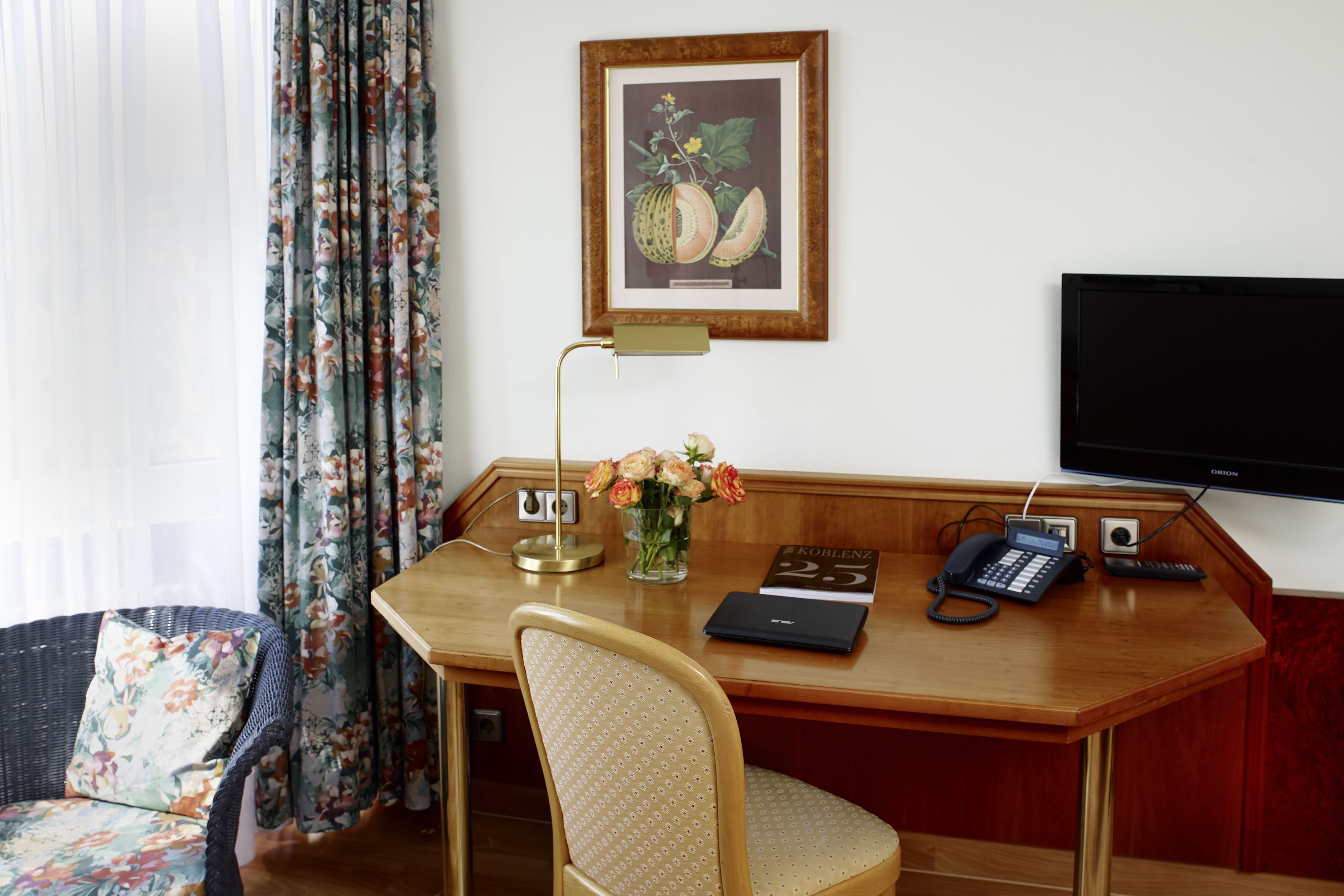 Une chambre d’hôtel avec un bureau en bois, une chaise, une estampe botanique encadrée, un vase avec des fleurs, une lampe de bureau, un téléphone et un téléviseur.