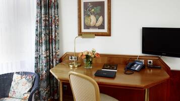 Une chambre d’hôtel avec un bureau en bois, une chaise, une estampe botanique encadrée, un vase avec des fleurs, une lampe de bureau, un téléphone et un téléviseur.