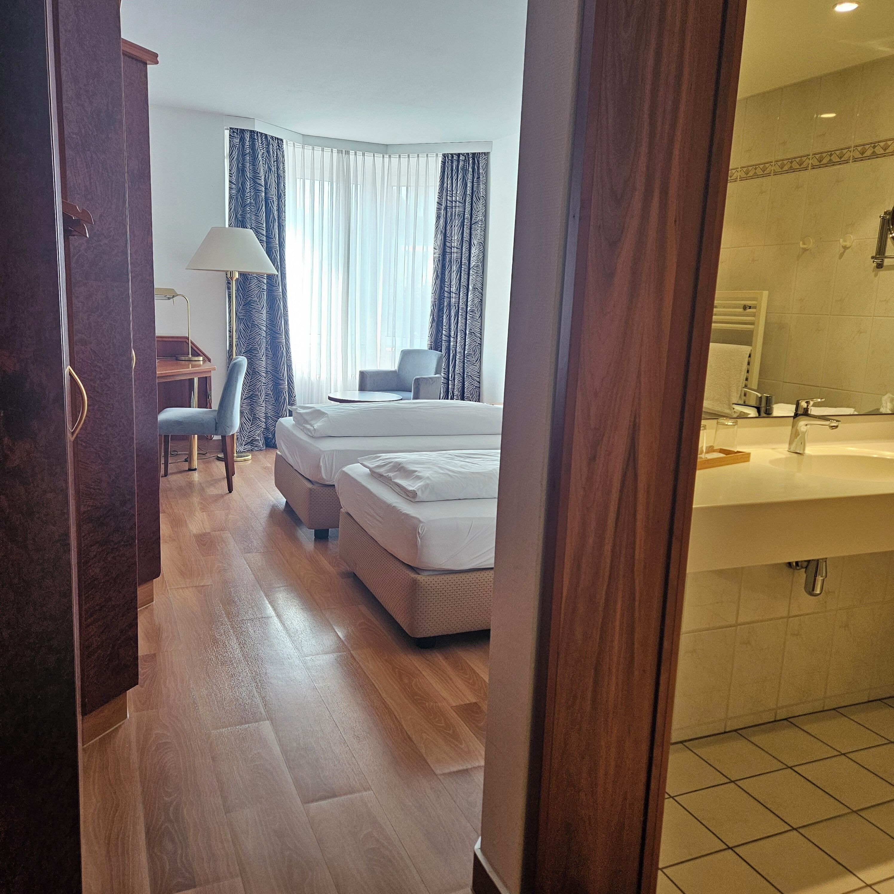 Habitación de hotel con cama, escritorio, silla, lámpara y baño con lavamanos.