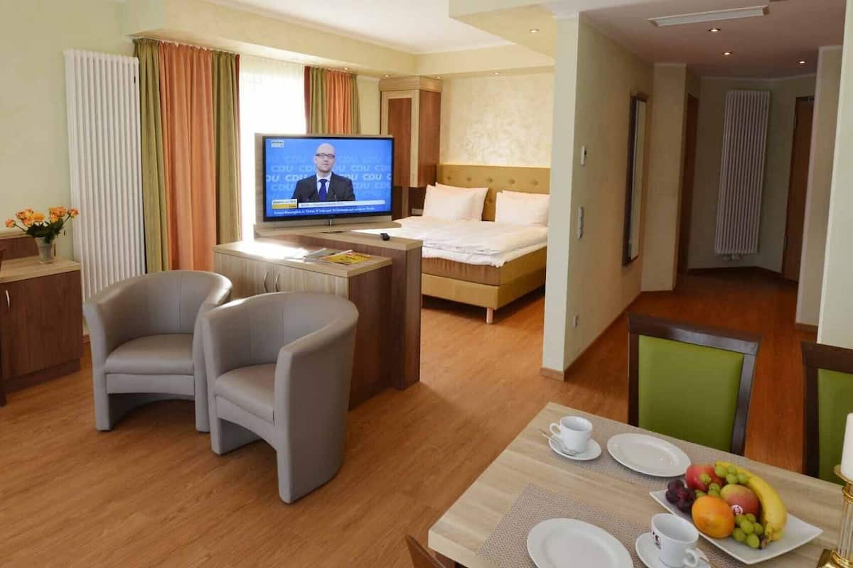 A hotel room with a bed, a desk with a television, two armchairs, a dining table with plates and a candle, and a window with curtains.