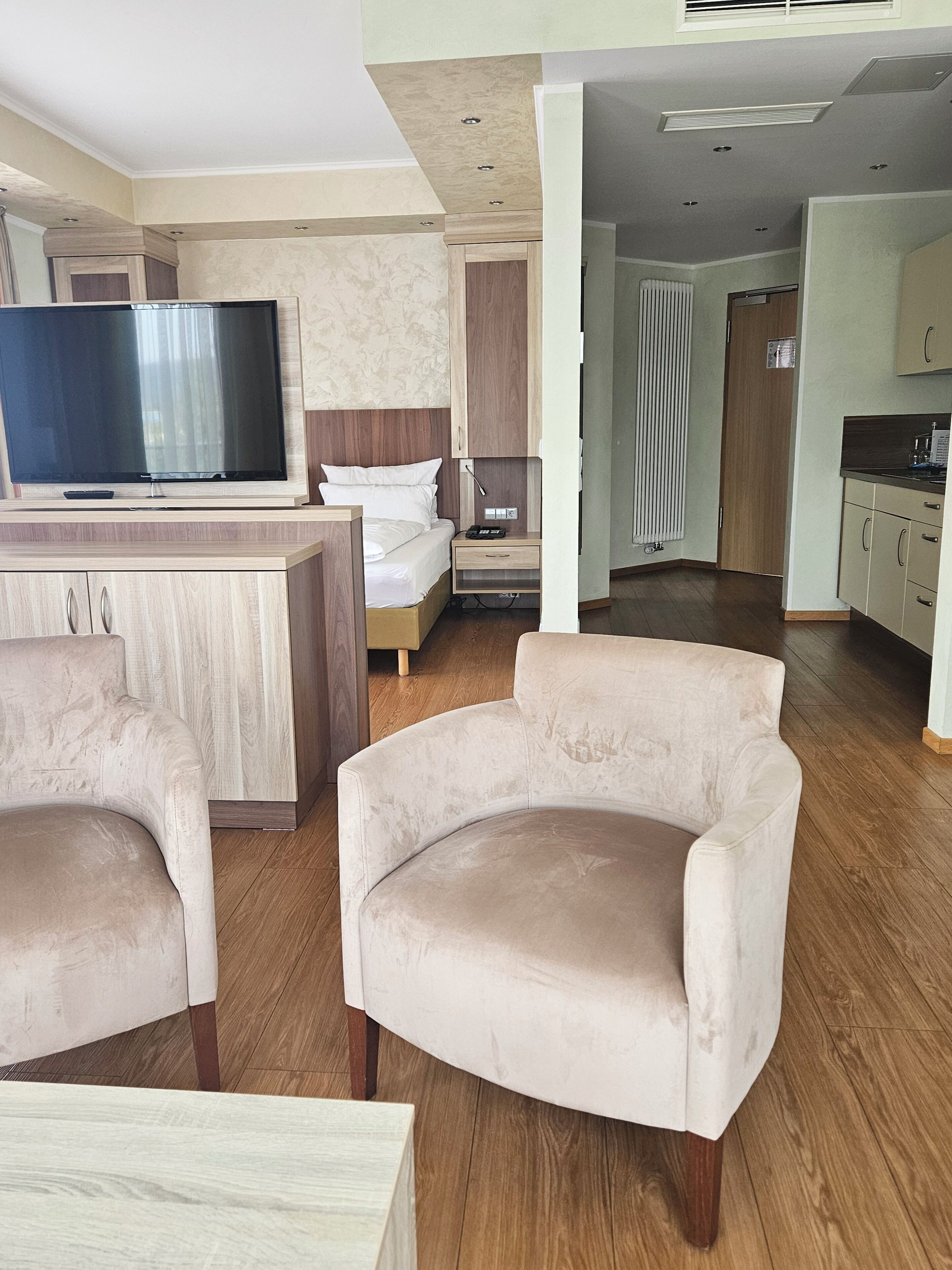 Habitación de hotel moderna con televisor de pantalla plana, cama con ropa blanca, escritorio de madera y zona de estar con dos sillones.