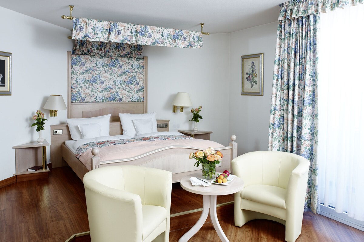 A hotel room with a large bed, two bedside tables with lamps, a small round table with a vase of flowers, two armchairs, and floral curtains.