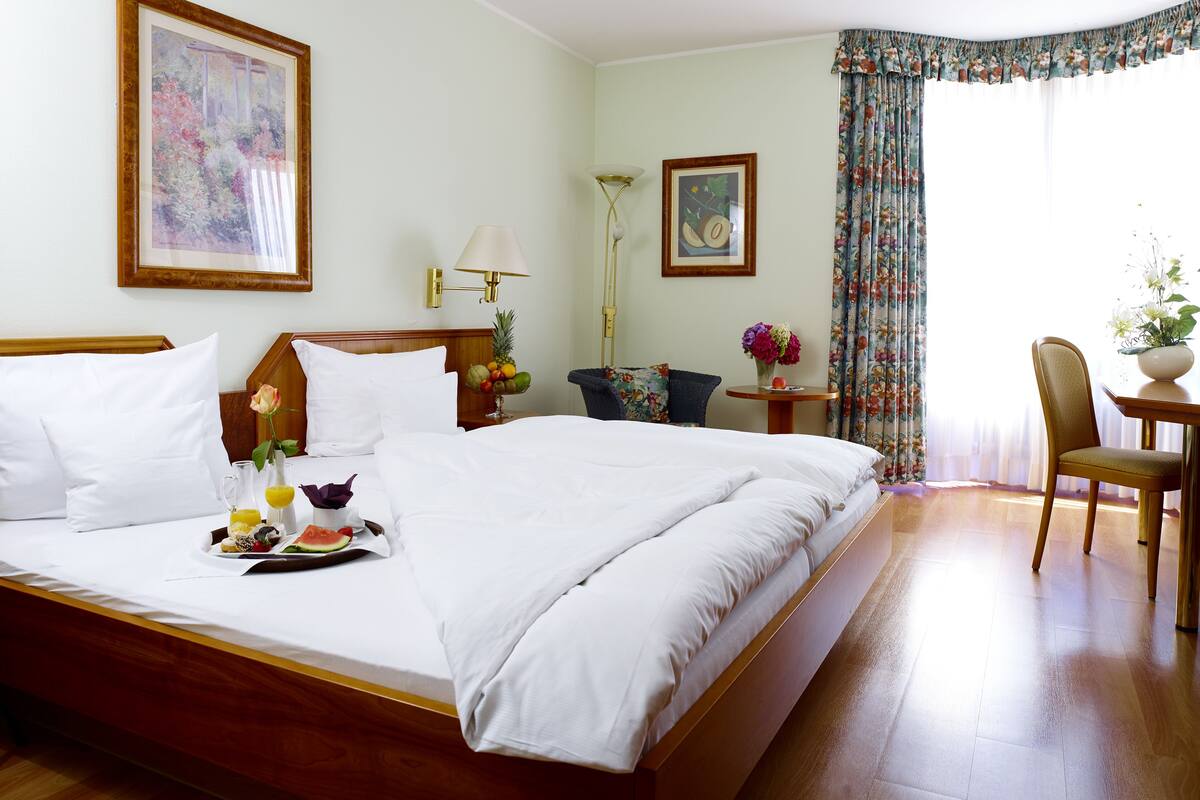 A hotel room with a large bed, a bedside table with a lamp, a chair, a small table with a vase of flowers, and a framed picture on the wall.