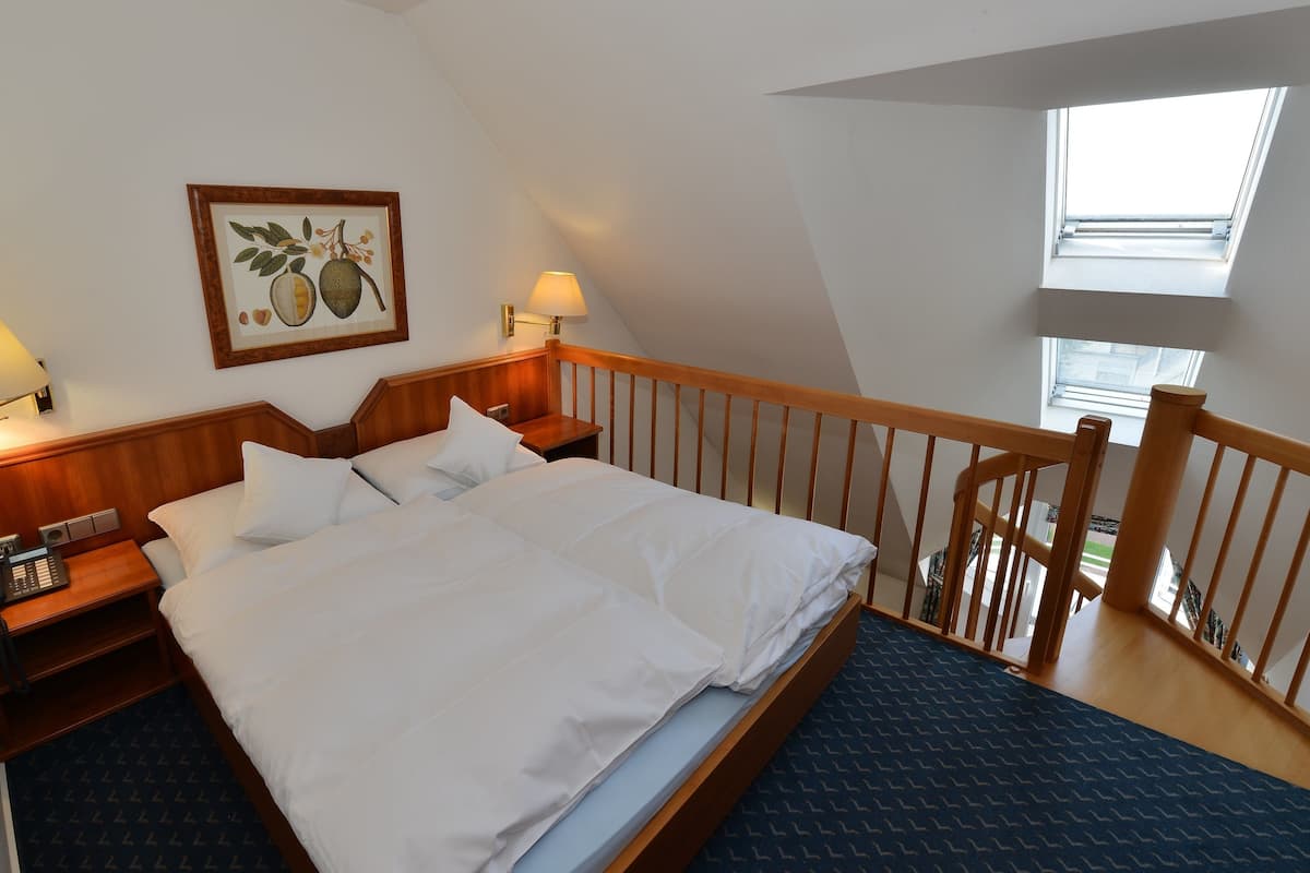 A bedroom with a large bed, wooden headboard, two bedside tables with lamps, a wall-mounted telephone, and a window with a view of the outside.