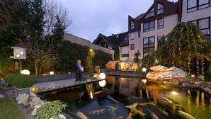 A koi pond with rocks and a birdhouse, surrounded by a garden and a building with a sloped roof. - Rheinhotel LARUS (Kaltenengers)