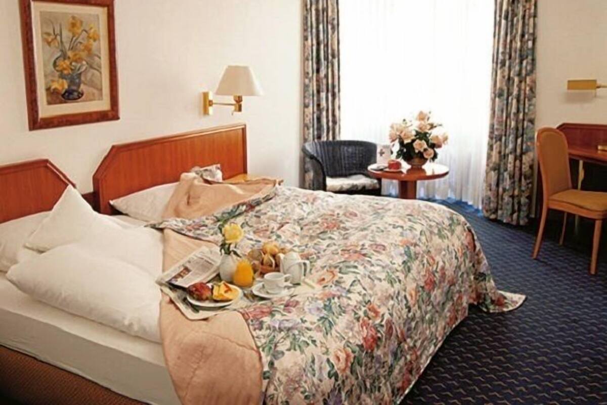 A hotel room with a bed, a chair, a table with a vase of flowers, and a painting on the wall.