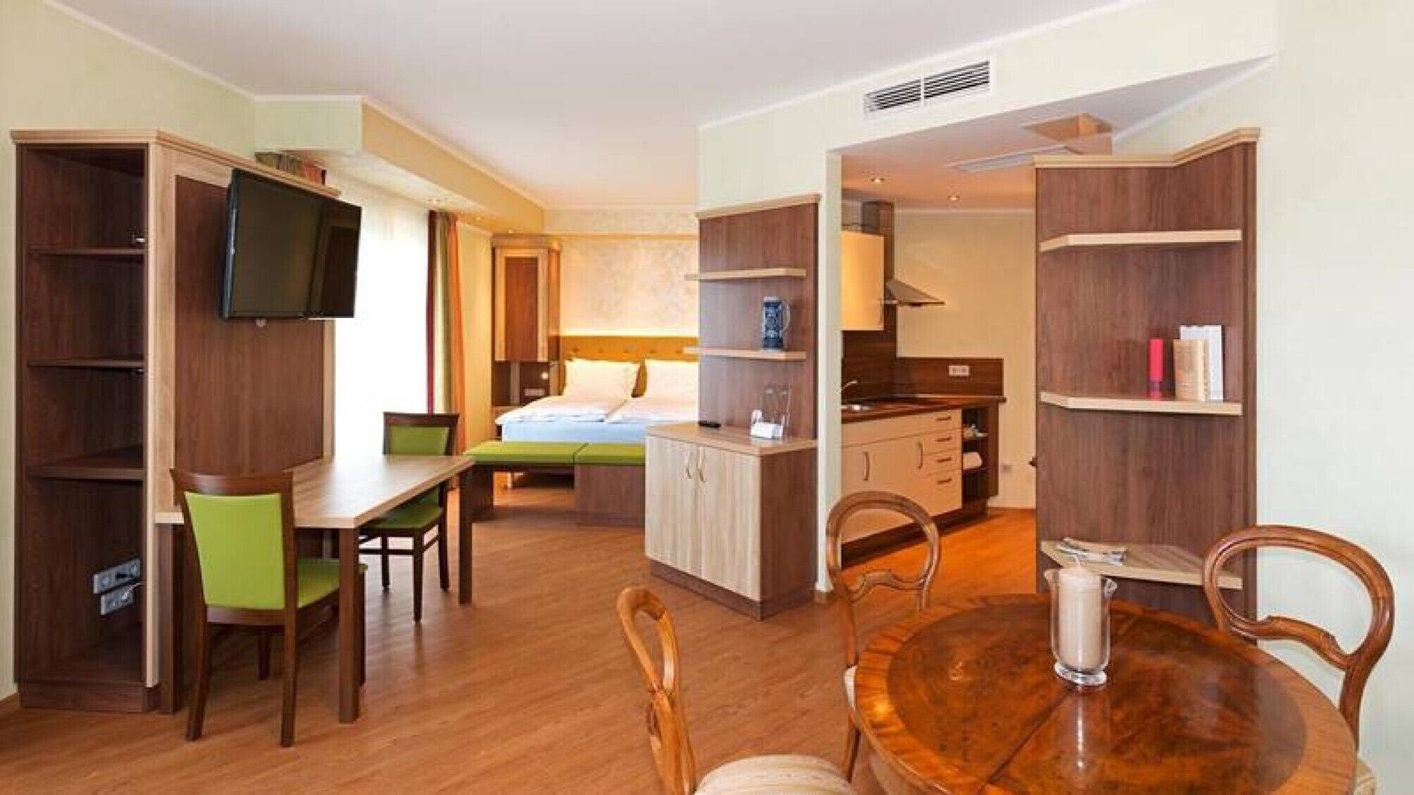 A compact hotel room with a bed, a desk, a chair, a small kitchenette, and a TV mounted on the wall.