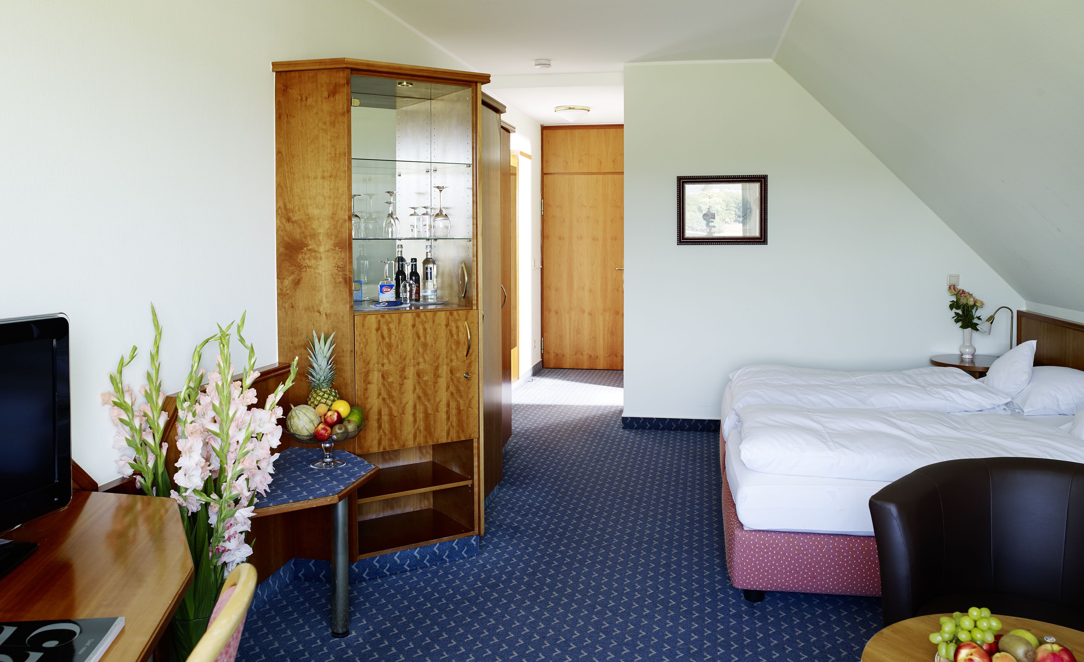A hotel room with a bed, a desk with a computer, a wardrobe, and a small table with flowers.