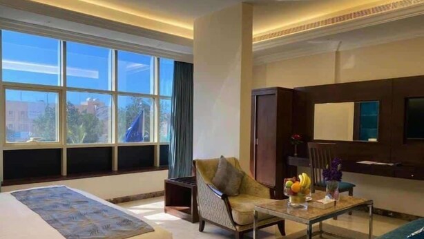 Habitación de hotel con una cama grande, un sofá, una mesita con frutas, un televisor y una ventana con vistas a edificios.