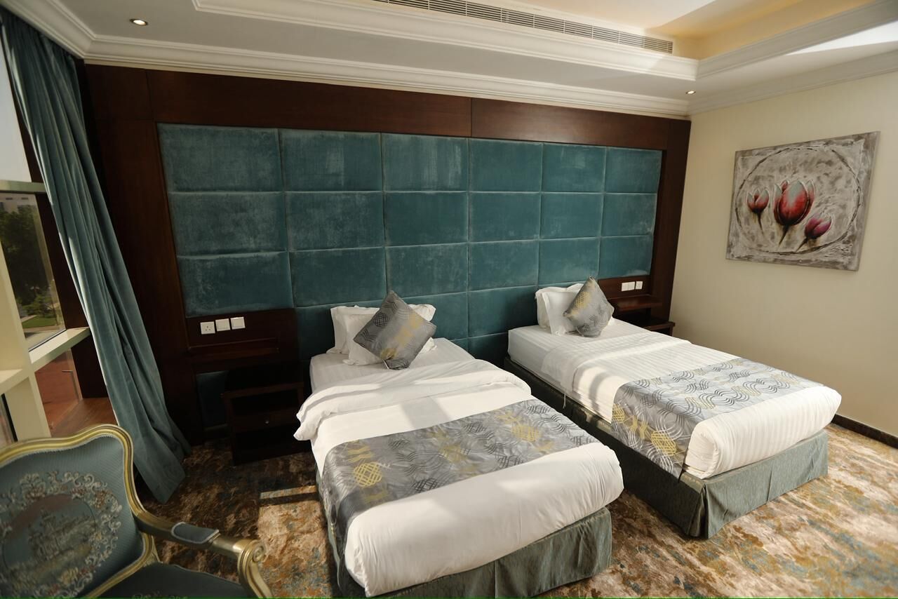 A hotel room with two beds, a large teal headboard, a painting of flowers, and a chair with a decorative backrest.