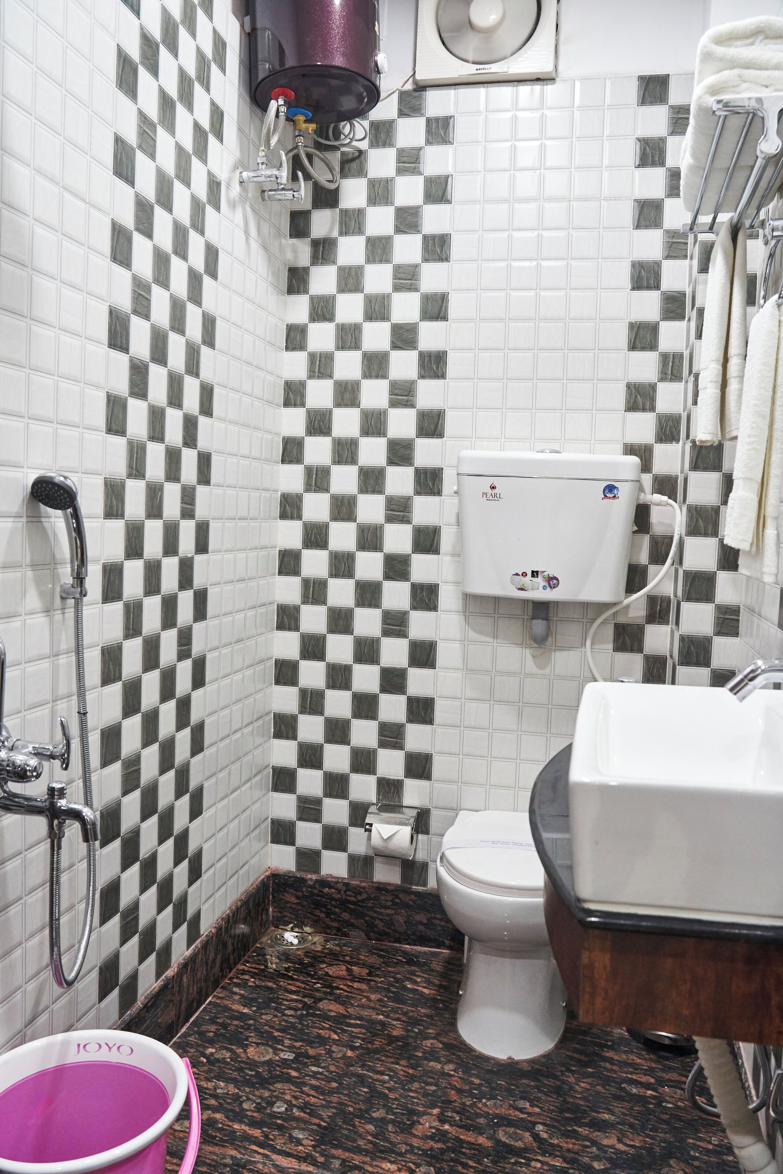 A bathroom with a checkered tile wall, a toilet, a sink, a shower, and a bucket labeled "JOYO".