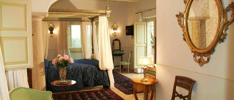 A bedroom with a four-poster bed, a green armchair, a wooden chair, a small table with a lamp, and a mirror on the wall.