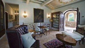 A spacious living room with a leather sofa, a wooden coffee table, and a patterned rug. There is a dining area with a table and chairs, and a large painting on the wall. The room has a high ceiling with decorative moldings and a view through an arched doorway to an outdoor area. - Relais Tenuta del Gallo (Amelia)