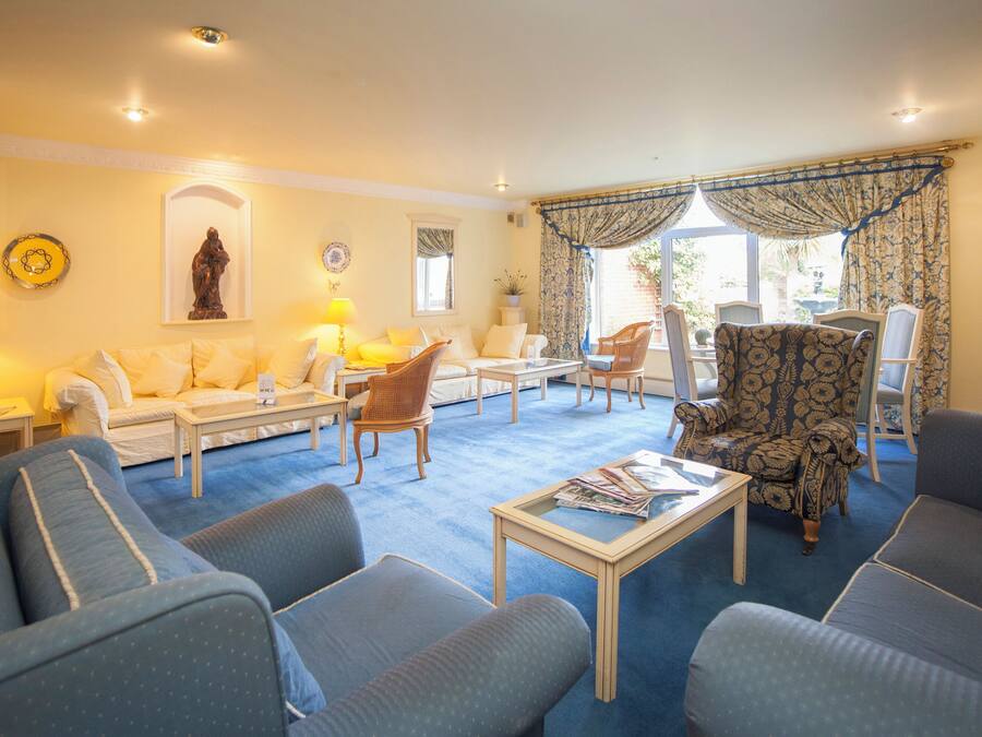 A well-lit living room with a blue carpet, white sofas, a patterned armchair, and a coffee table.