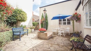 A patio area with stone flooring, a blue bench, white chairs, a table, and hanging flower baskets. - The Walnut Tree Hotel (Bridgwater)