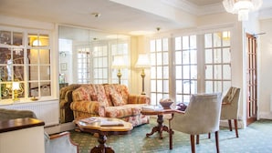 A cozy living room with a floral sofa, a wooden coffee table, and a chair. There are large windows with white frames and a chandelier. - The Walnut Tree Hotel (Bridgwater)