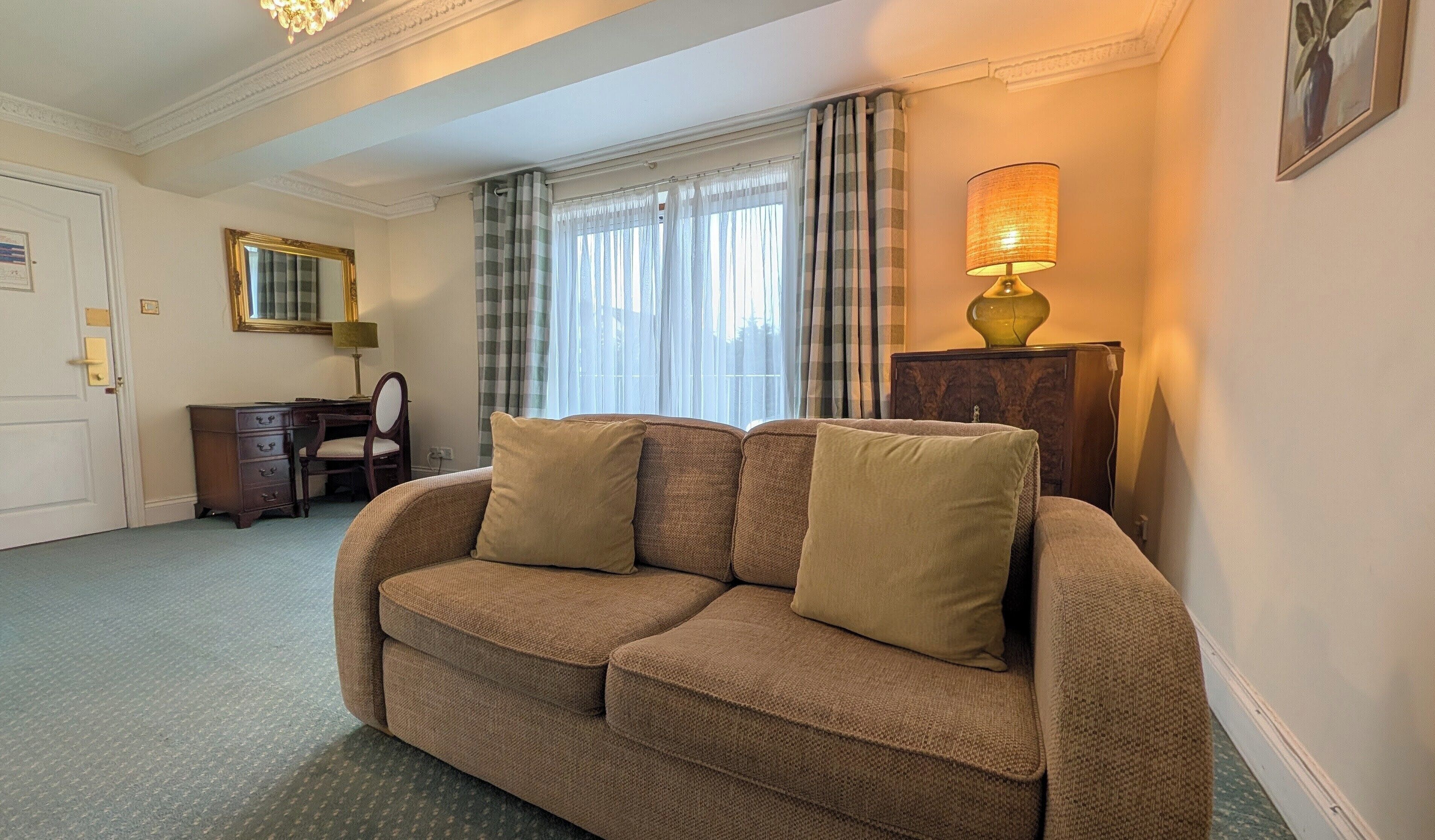 Habitación de hotel con un sofá marrón, un escritorio con una silla, una lámpara y una ventana con cortinas.