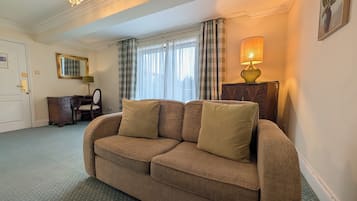 Habitación de hotel con un sofá marrón, un escritorio con una silla, una lámpara y una ventana con cortinas.
