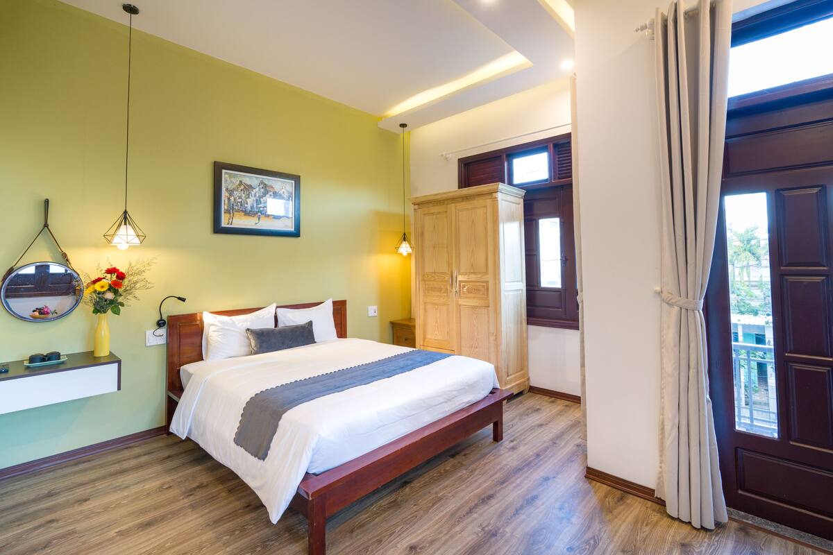 Ein Schlafzimmer mit einem hölzernen Bett, einem Nachttisch, einem Spiegel, einem an der Wand befestigten Regal mit einer Vase und einem Fenster mit Vorhängen.