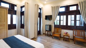 A hotel room with a bed, a television, two wooden chairs, a small table with a flower, and large windows with curtains. - Le Printemps D' An Bang (Da Nang)