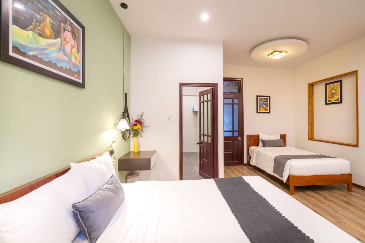 Ein Hotelzimmer mit zwei Einzelbetten, einem Nachttisch mit Blumenvase, einem Bild an der Wand und einer Tür zu einem weiteren Raum.