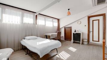 A hotel room with a bed, a desk, a chair, a small table with a kettle, a clock, and a door.