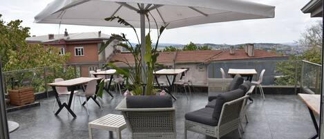 Un patio al aire libre con mesas y sillas, un paraguas grande y vista panorámica de la ciudad.