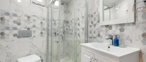 Un baño moderno con ducha envuelta en vidrio, azulejos de mármol blanco y un tocador blanco con espejo.