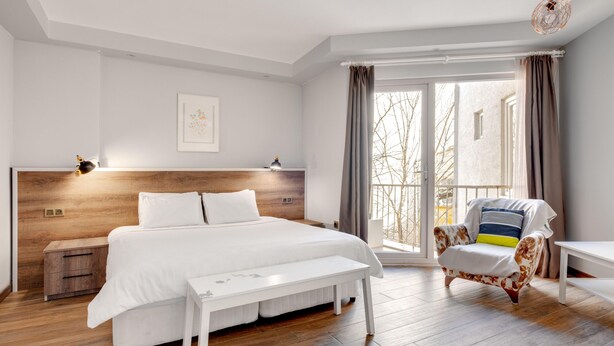 A modern bedroom with a wooden headboard, a large bed, a bench, a chair, and a view of the outdoors through a sliding door. - Yankı Hotel (Bursa)
