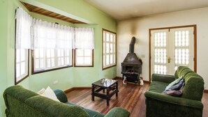 Uma sala de estar com um sofá verde, uma mesa de centro de madeira, cortinas de renda e uma lareira a lenha.