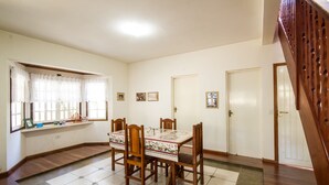 Uma sala de jantar com uma mesa posta para quatro pessoas, rodeada por cadeiras de madeira, piso ladrilhado e uma janela com cortinas.
