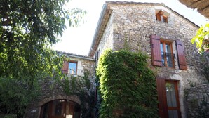A stone building with a wooden door and window shutters, surrounded by greenery and a stone archway.