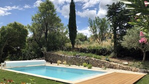 A rectangular swimming pool with a wooden deck in the foreground, surrounded by a stone wall and lush greenery.