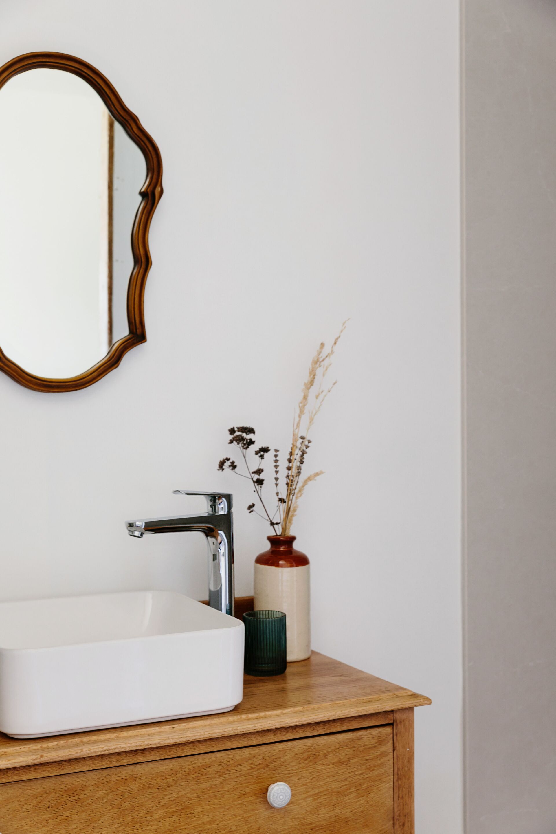 A bathroom with a white sink, a wooden vanity, a mirror with a wooden frame, and a vase with dried flowers.