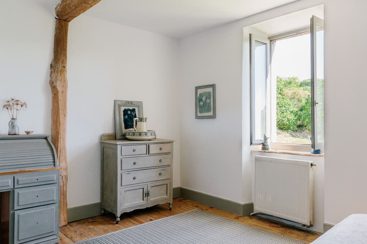 A room with a wooden floor, a dresser with drawers, a radiator, and a window with a view of greenery.