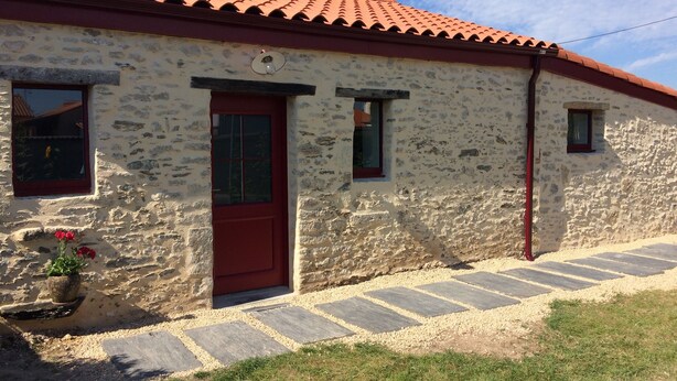 Ein Steinhaus mit roter Tür und Fenstern, ein kleiner Garten mit Blumen und ein Ziegeldach.