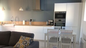 Eine moderne Küche mit weißen Schränken, einem eingebauten Backofen und einer Kaffeemaschine. Es gibt einen Esstisch mit weißen Stühlen und ein graues Sofa mit gemusterten Kissen.