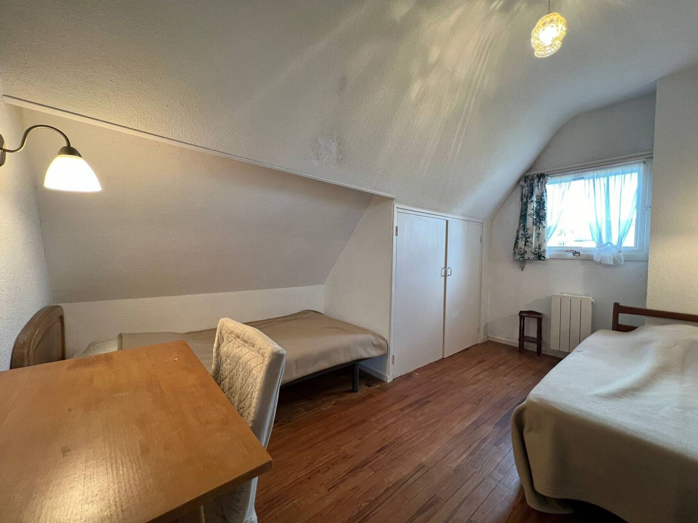 Una piccola e accogliente stanza mansardata con un tavolo di legno, un letto singolo e una finestra con tende.