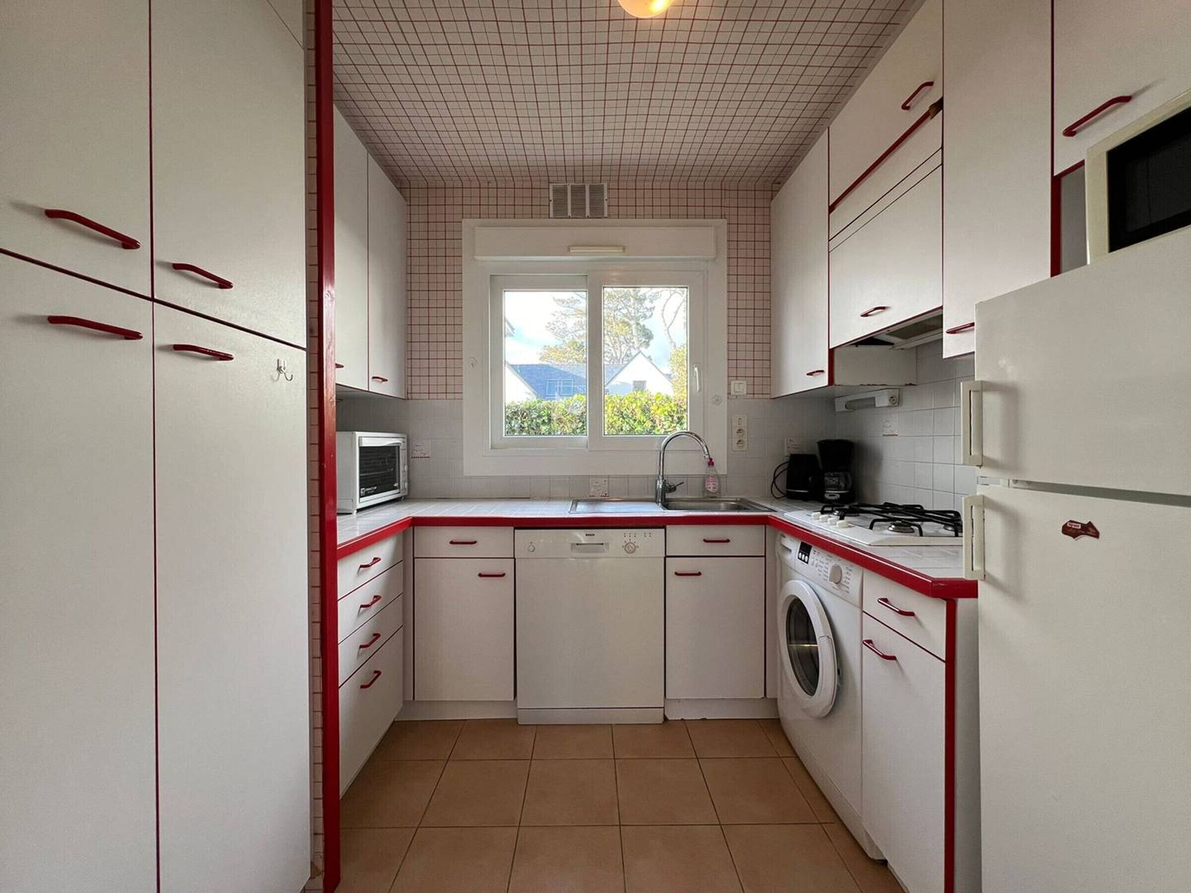 A compact kitchen with white cabinets, red handles, a built-in washing machine, and a window providing natural light.