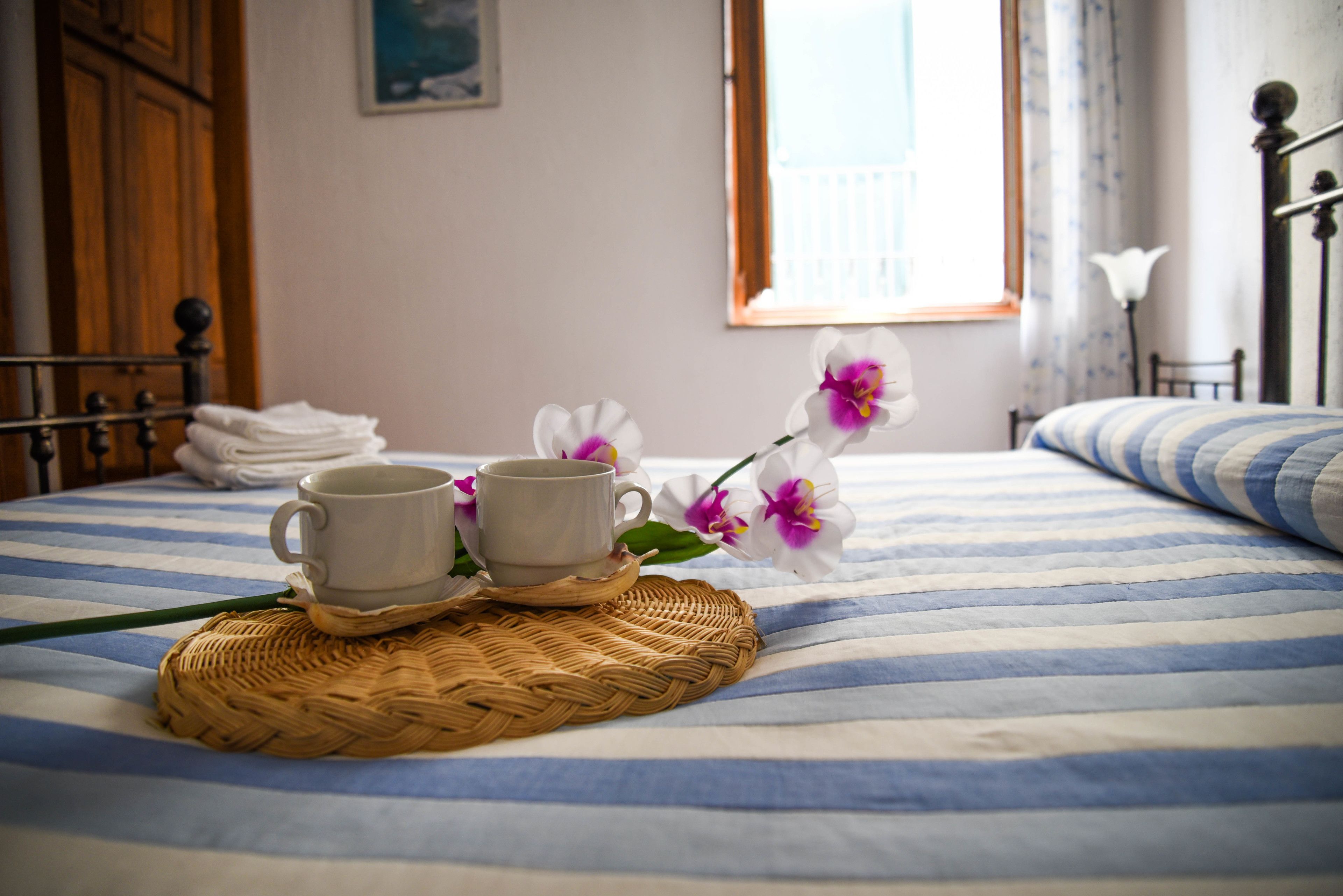 Una cama con colcha a rayas, dos tazas blancas sobre un posavasos tejido, y una orquídea morada y blanca.