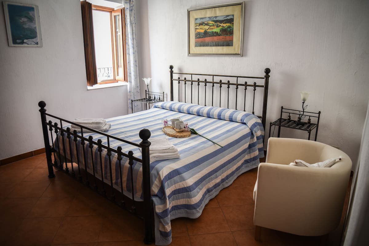 A bedroom with a bed, a chair, a nightstand, a window, and a painting on the wall.