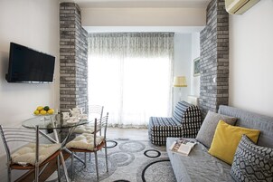 A modern living room with a glass dining table, metal chairs, a grey sofa with patterned cushions, and a brick accent wall.