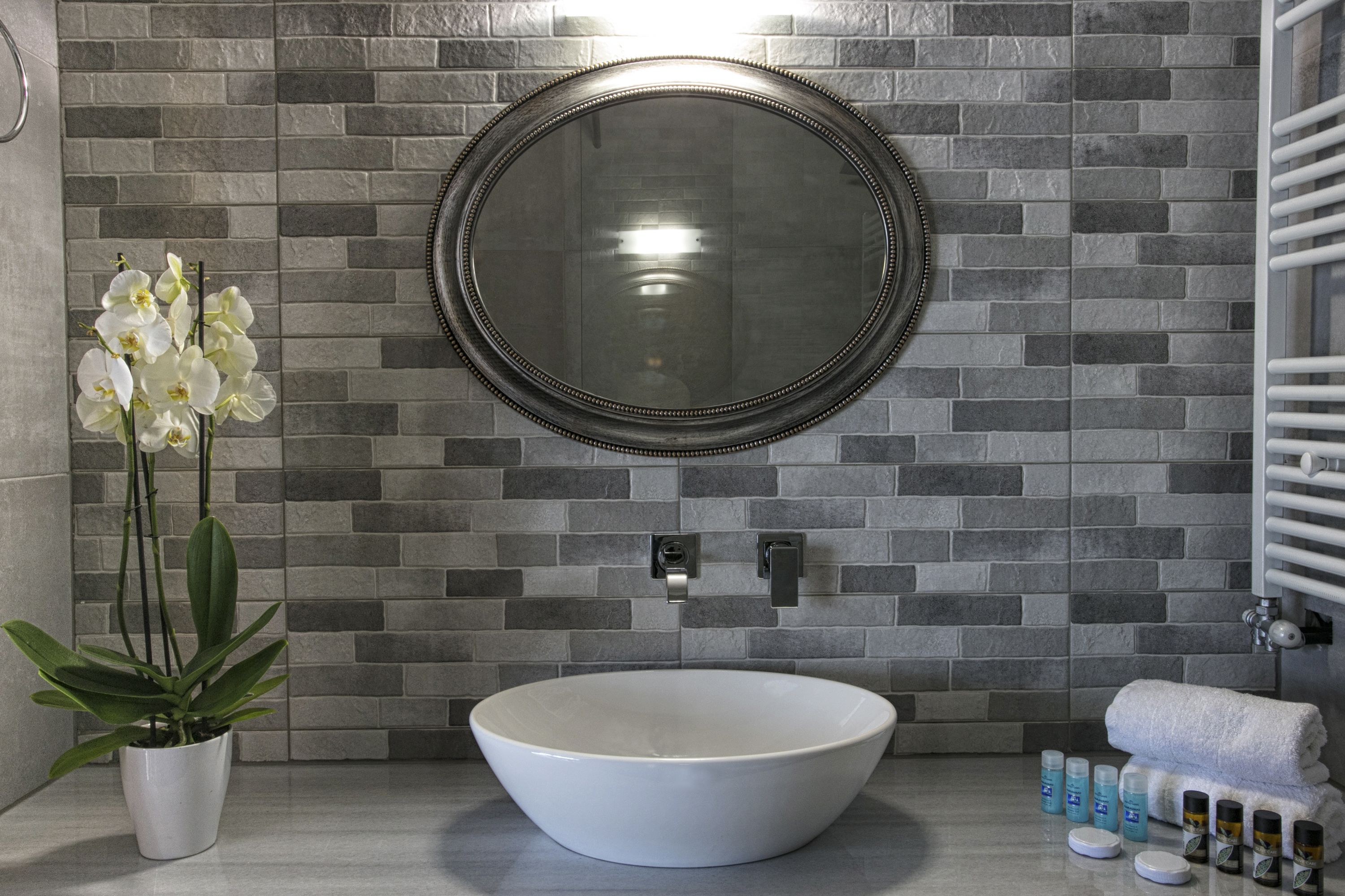 A bathroom with a white vessel sink, a round mirror, a potted orchid, and toiletries on the counter.