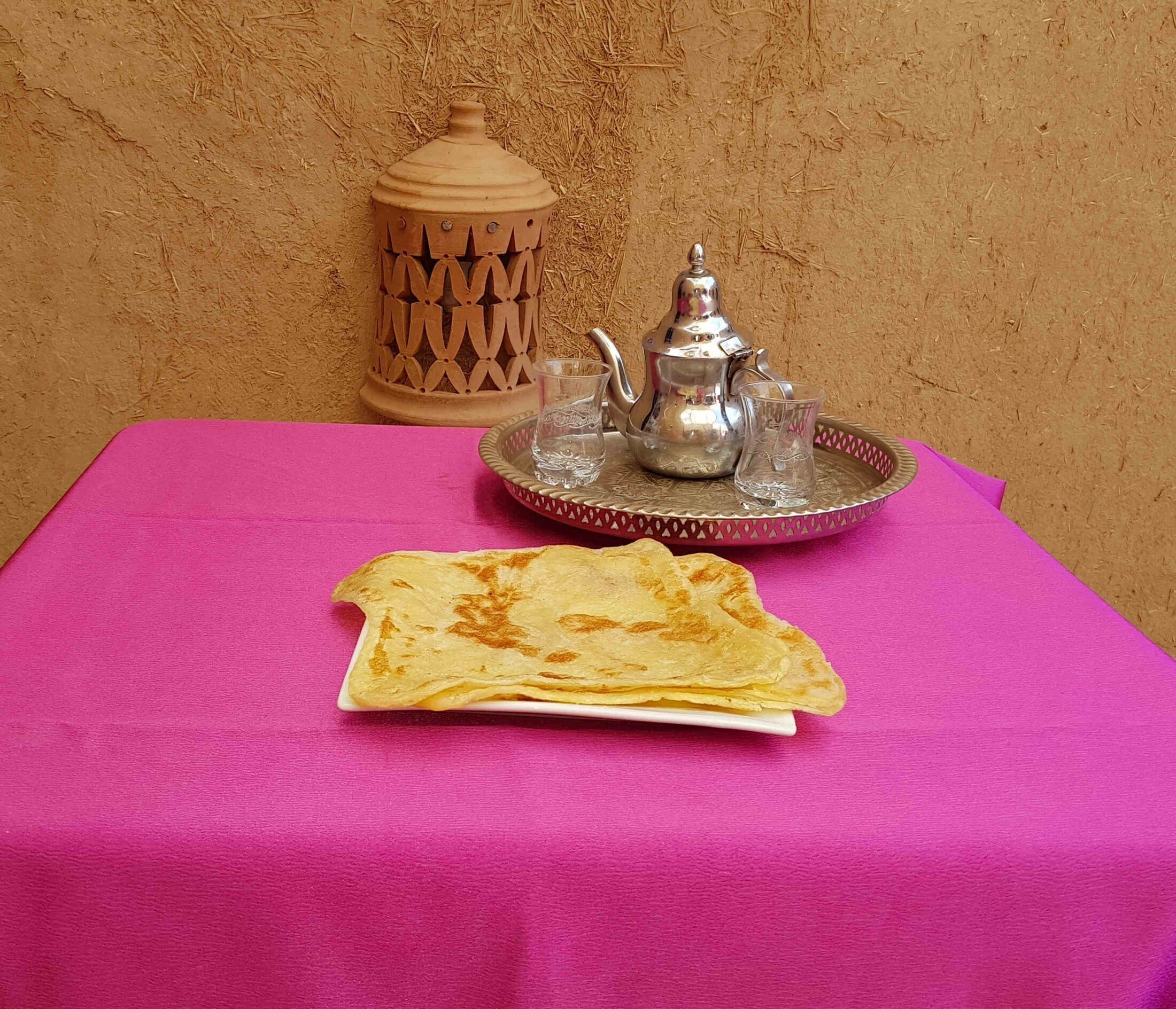 A table with a pink tablecloth, a silver teapot, two glasses, and a folded piece of bread.